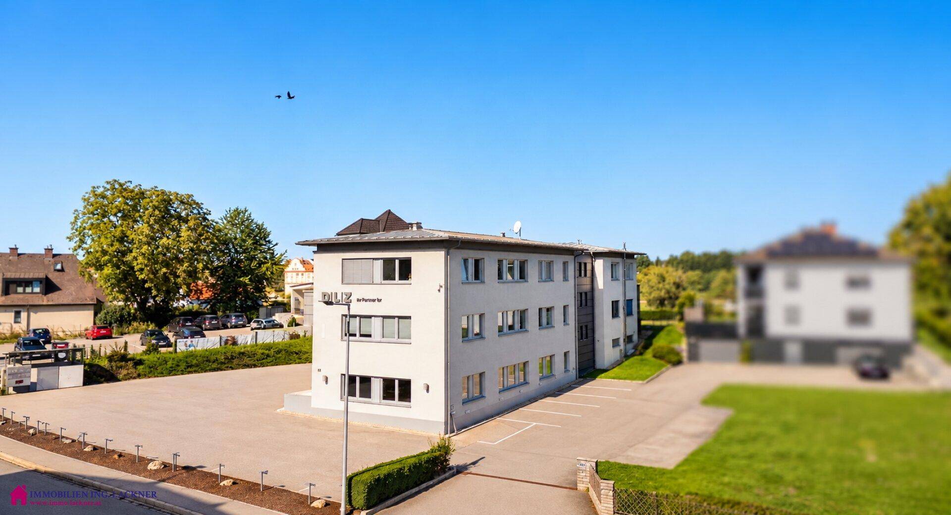 Luftaufnahme des modernen Bürogebäudes mit umliegenden Parkplätzen und Grünflächen unter blauem Himmel.
