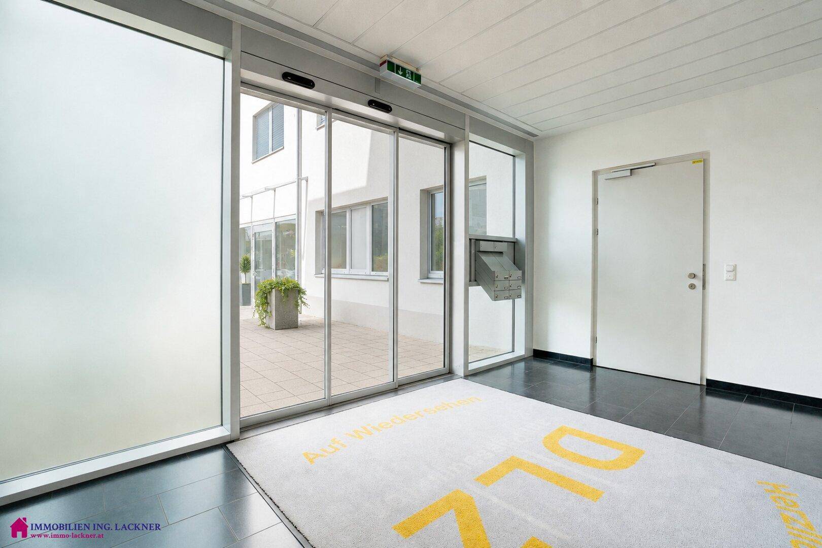 Heller Eingangsbereich mit automatischen Glastüren, dunklem Fliesenboden und Fußmatte.
