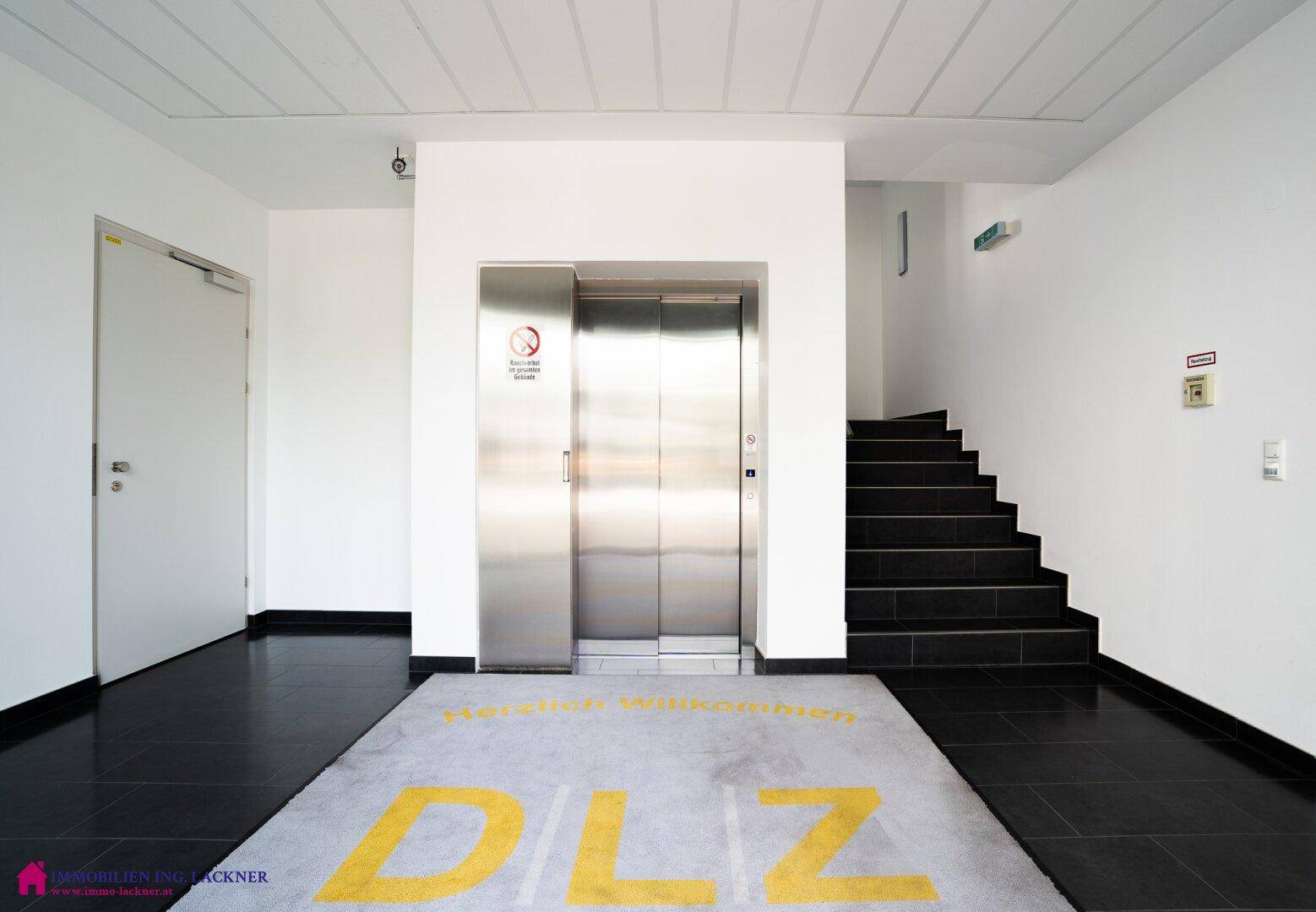 Heller Eingangsbereich mit Edelstahlaufzug, Treppenhaus und Willkommensmatte.