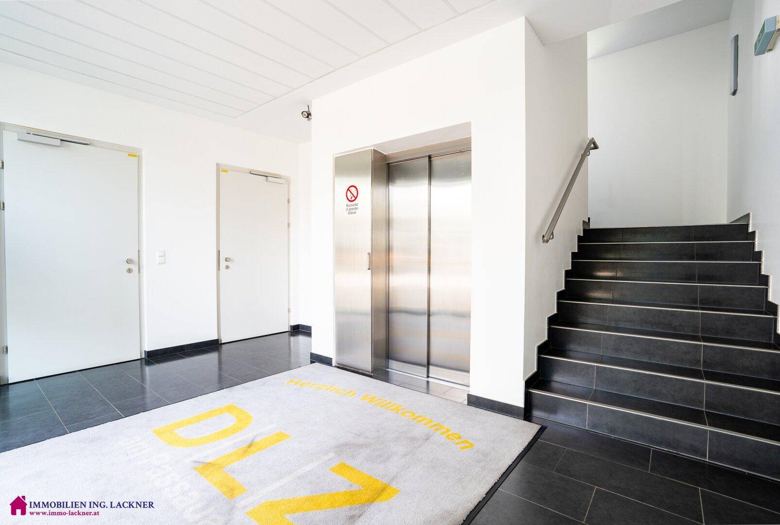 Moderner Eingangsbereich mit Aufzug, Treppe und dunklem Fliesenboden.