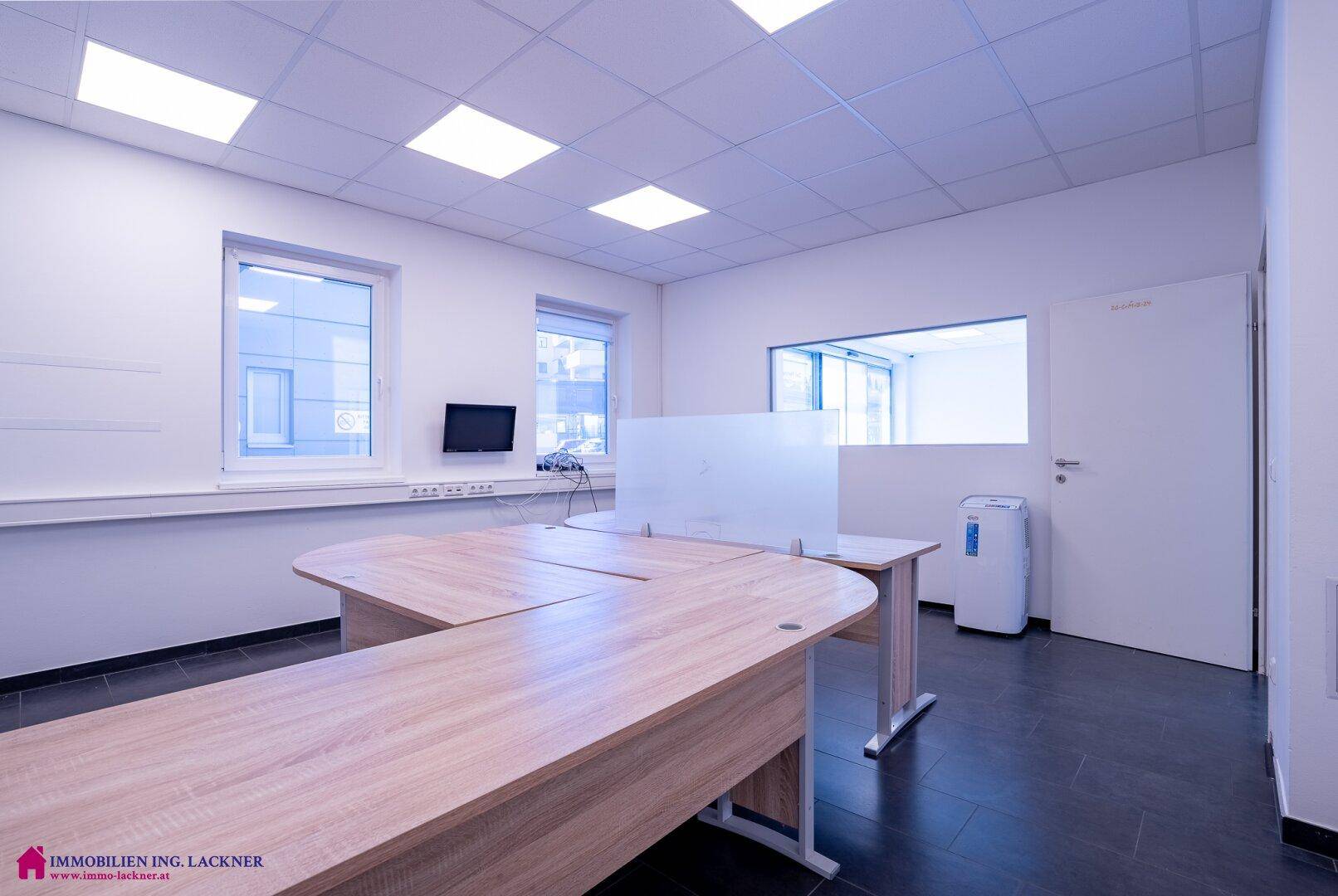 Modern eingerichtetes Büro mit hellen Holzschreibtischen, dunklem Fliesenboden und Fenstern.