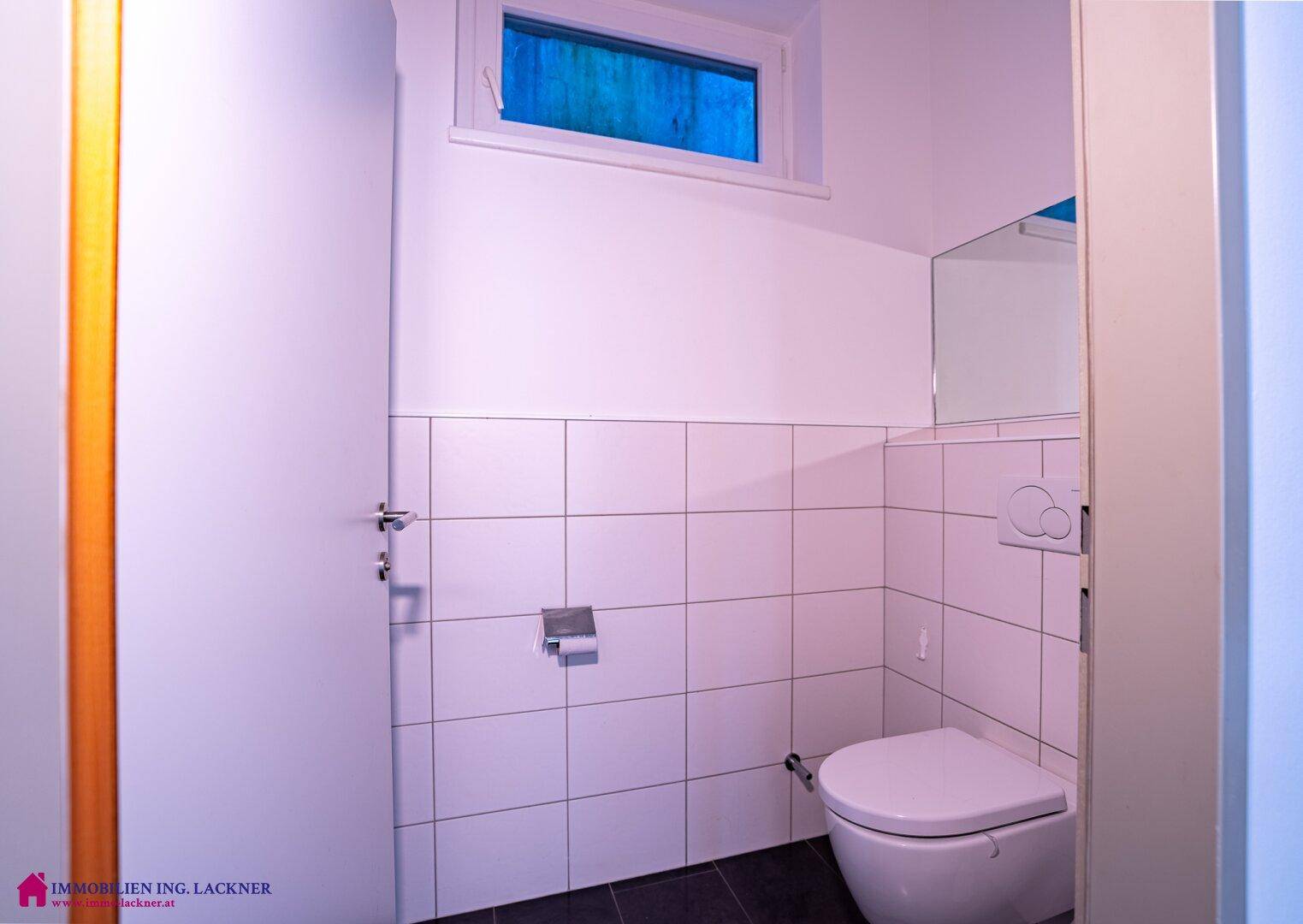 Saubere Toilette mit weißen Fliesen, wandmontiertem WC und kleinem Fenster für Belüftung.