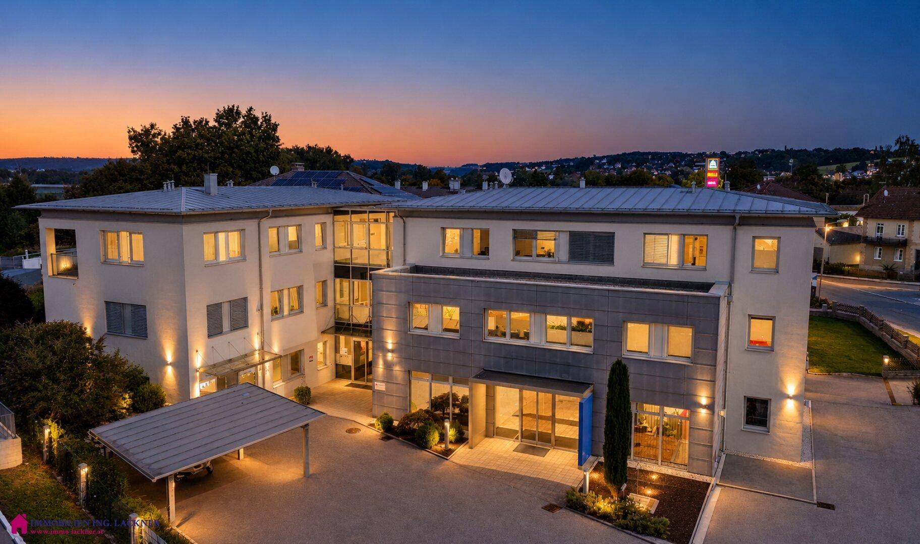 Ansicht des Bürokomplexes in der Abenddämmerung mit beleuchteten Fenstern und Außenbeleuchtung.