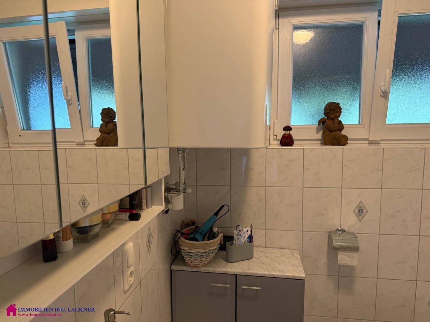 Badezimmer mit hellen Fliesen, Spiegelschrank und zwei Fenstern für Tageslicht.