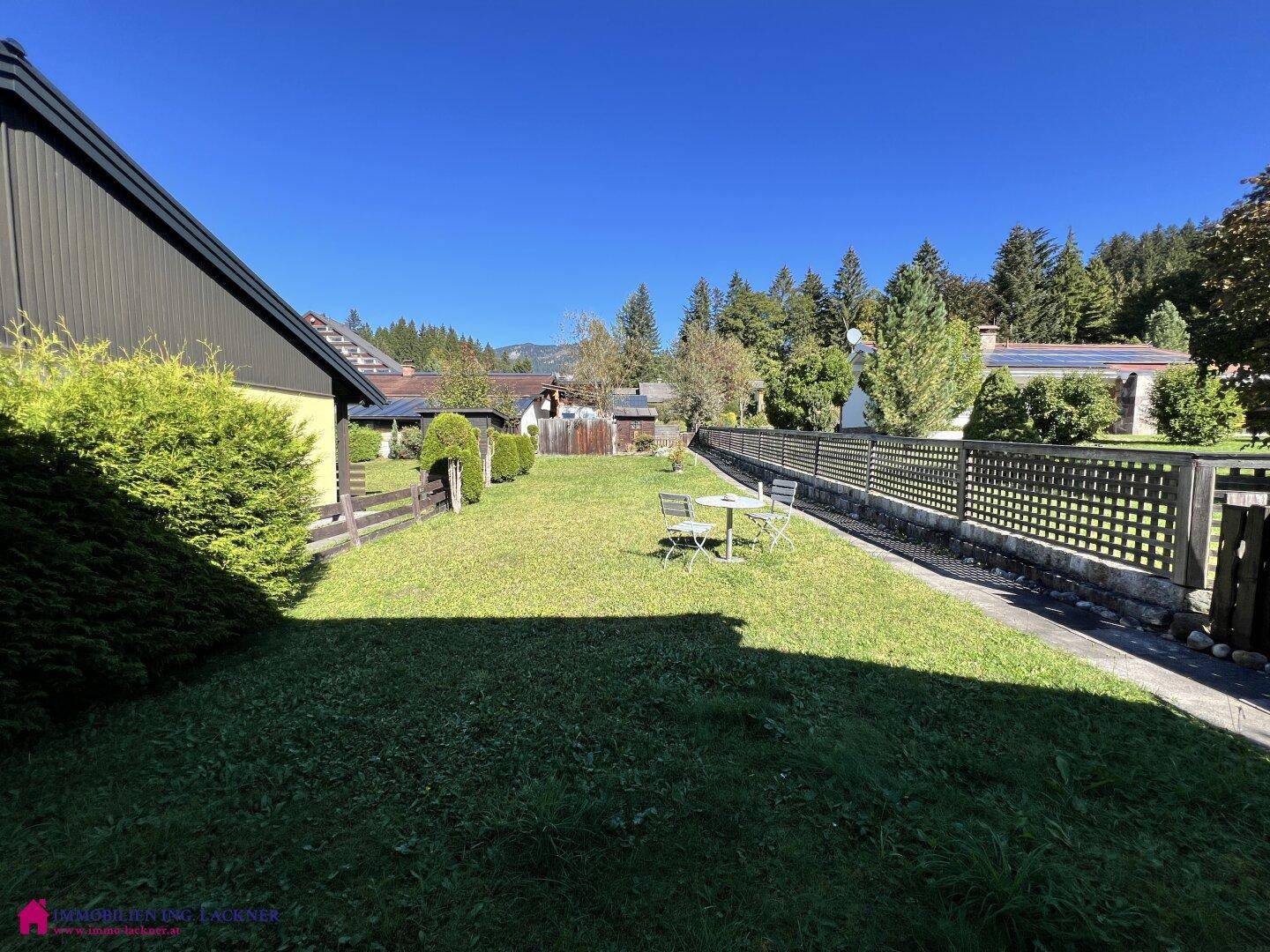 Langer, gepflegter Gartenbereich mit Rasen, Zaun und Sitzgelegenheit unter blauem Himmel.