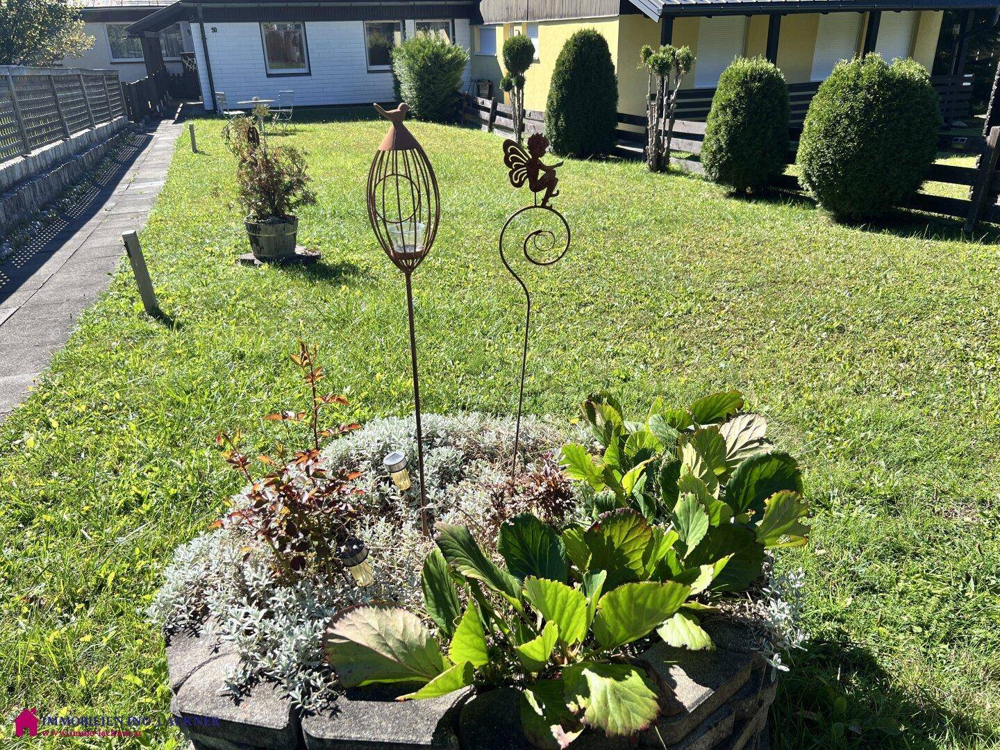 Grüner Garten mit Blumenbeet und dekorativen Elementen, umgeben von Rasenflächen.