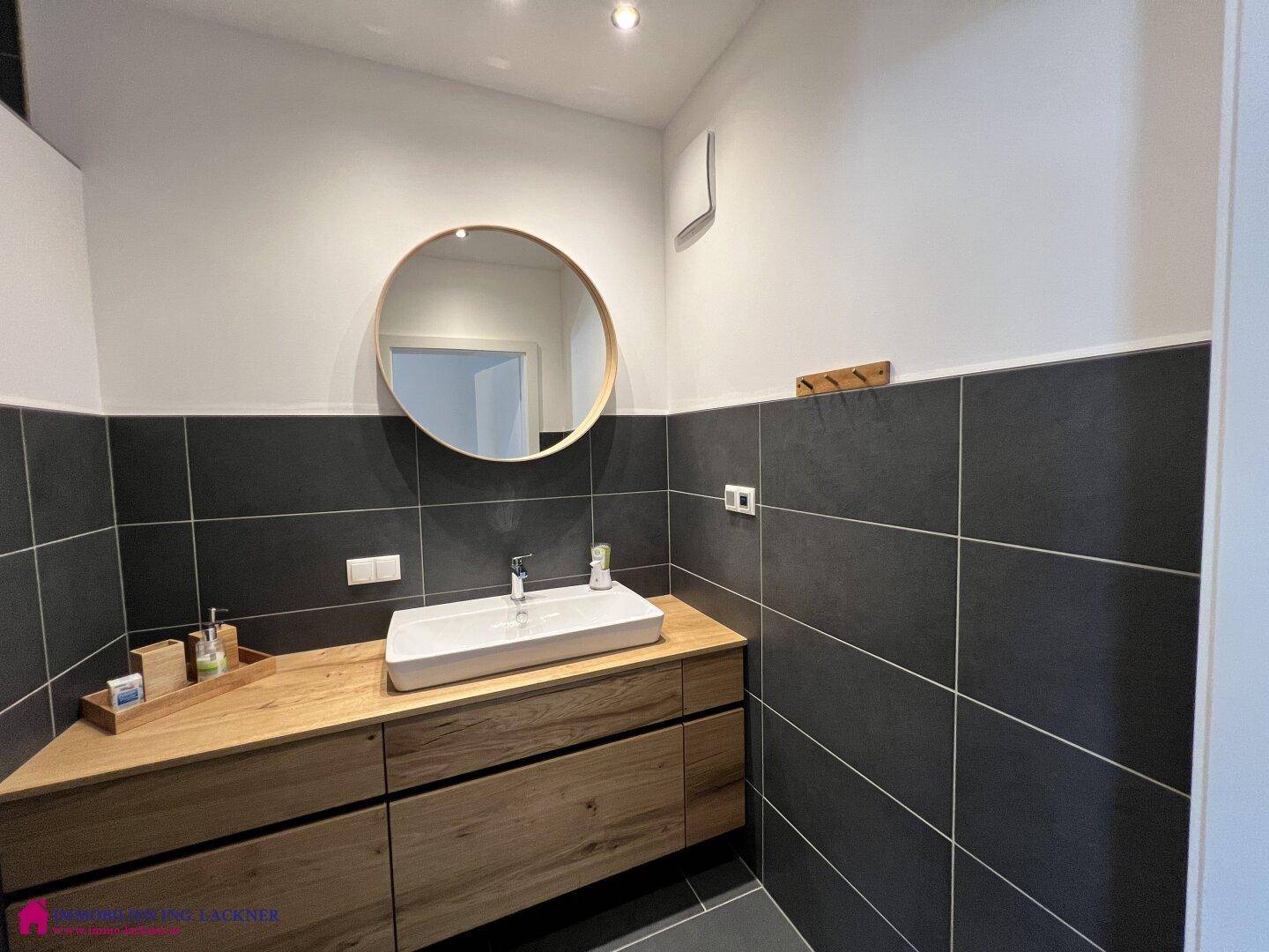 Modernes Badezimmer mit Holzwaschtisch, rundem Spiegel und dunklen Wand- und Bodenfliesen.
