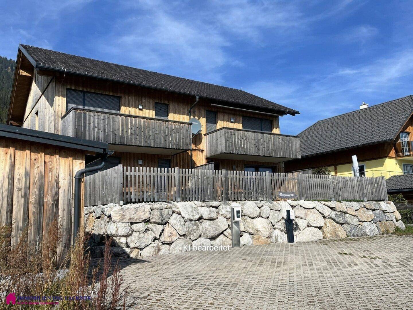 Vorderansicht eines Holzhauses mit Balkonen, Steinfassade und gepflastertem Parkplatz.