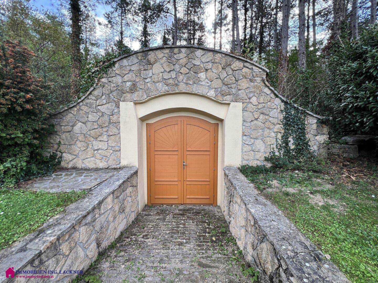 Rustikaler Eingang zu einem Weinkeller, eingebettet in eine Steinmauer und umgeben von Natur.