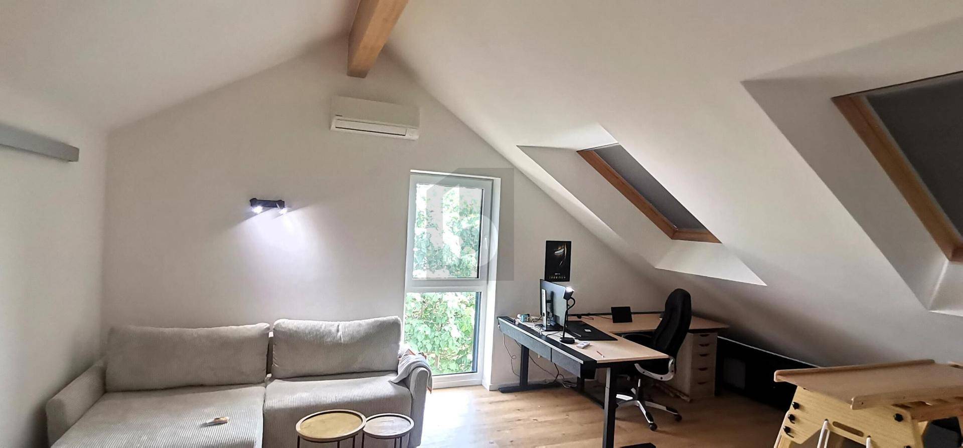 Helles Dachzimmer mit schrägen Decken, gemütlichem Sofa und Arbeitsbereich unter einem Dachfenster.