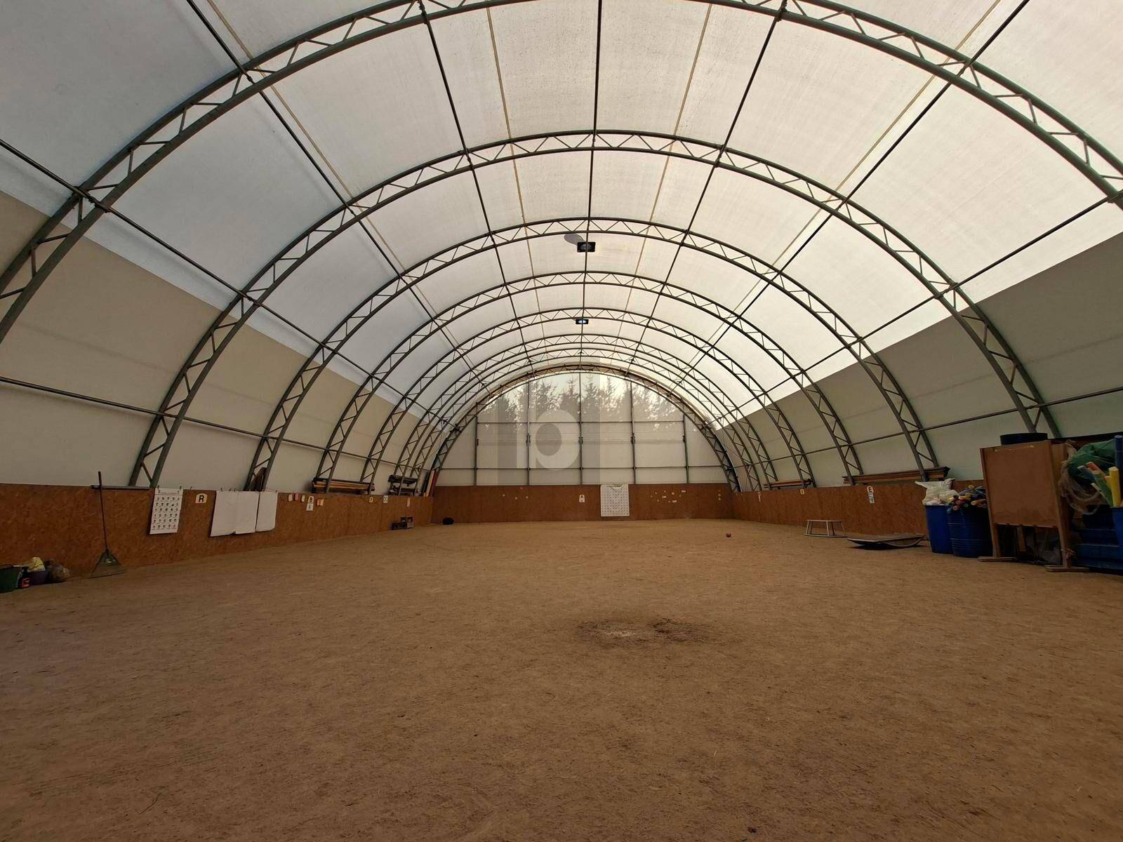 Große, bogenförmige Halle mit Sandboden und Metallkonstruktion, möglicherweise eine Reithalle oder Mehrzweckhalle.