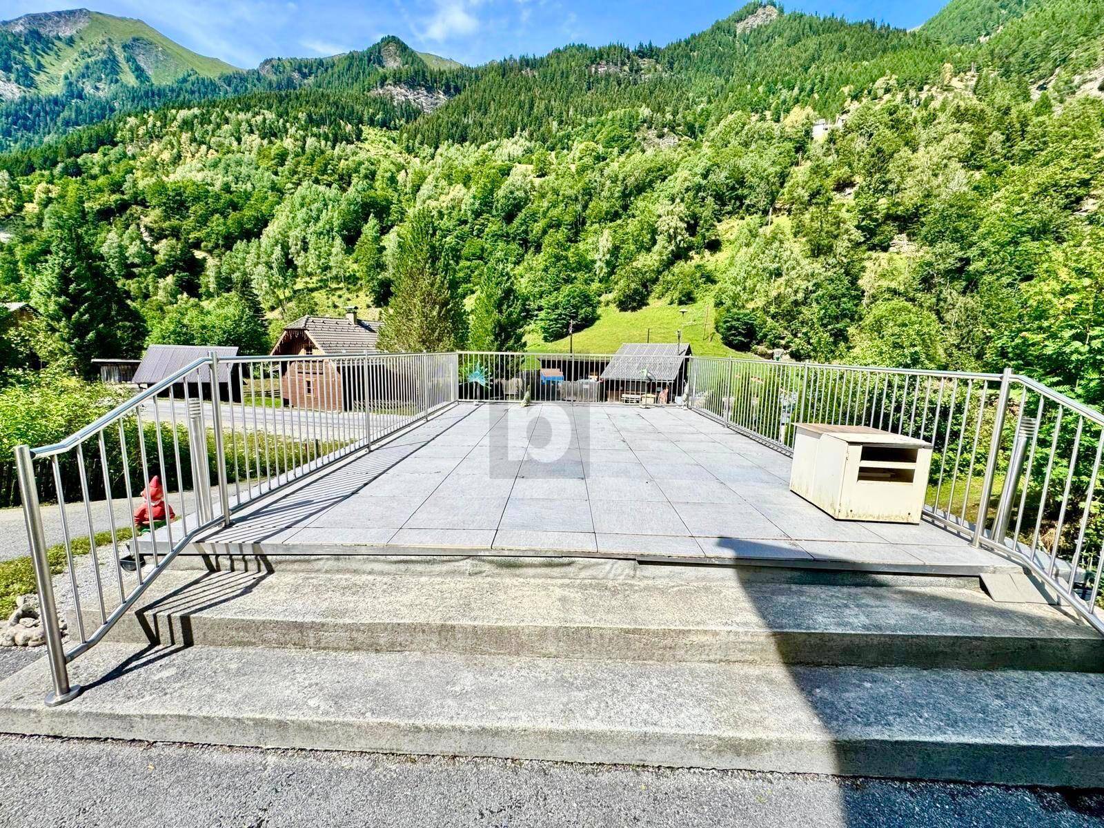 Große gepflasterte Terrasse mit Metallgeländer und beeindruckendem Blick auf die umliegenden grünen Berge.