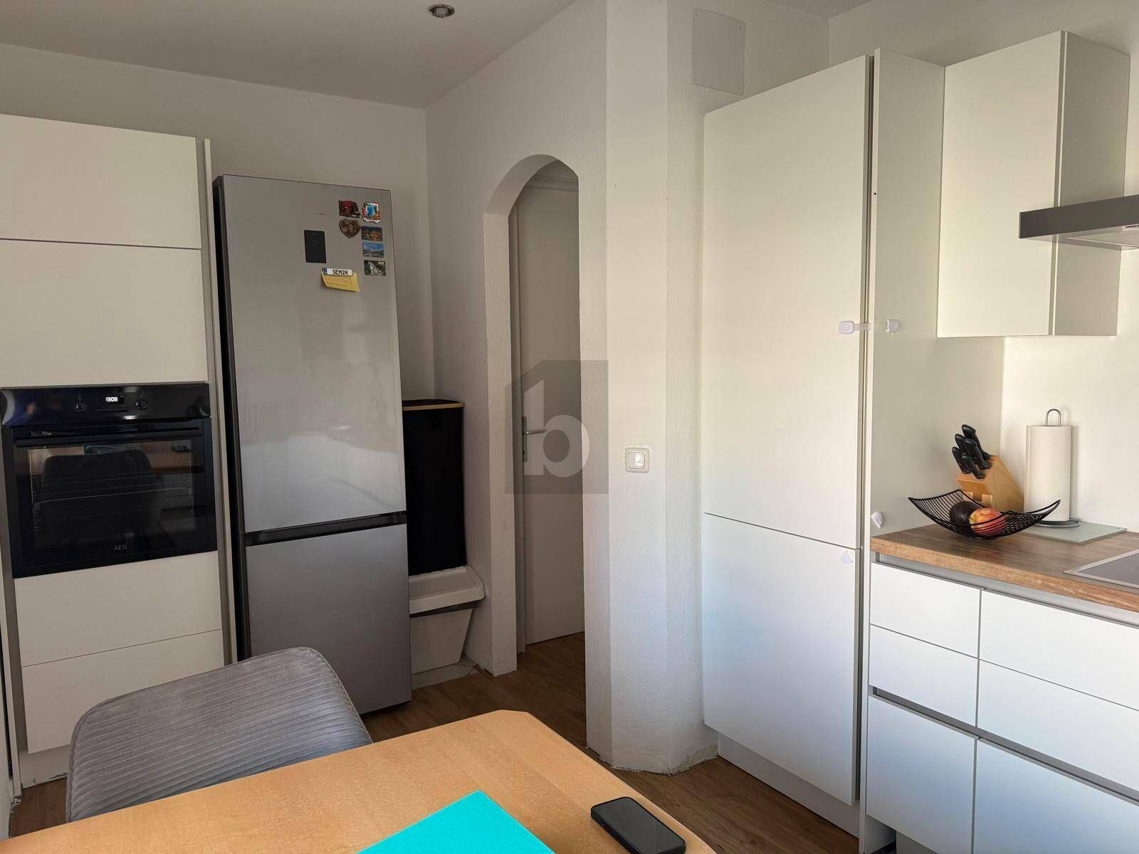 Küchenbereich mit Kühlschrank, Backofen, weißen Schränken und einem Holztisch.