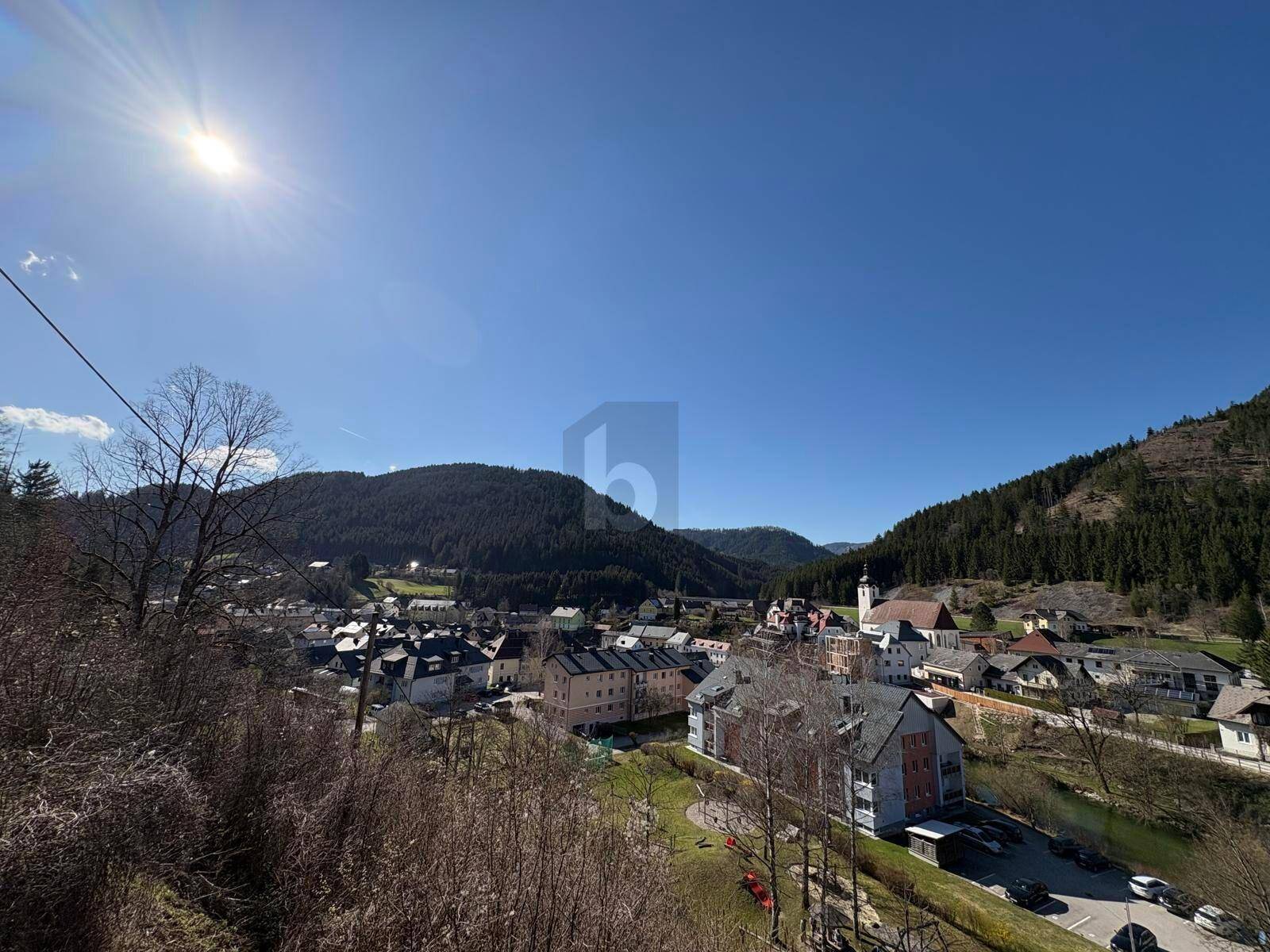 Panoramablick auf eine Stadt im Tal, umgeben von Bergen und Wäldern unter strahlendem Sonnenschein.