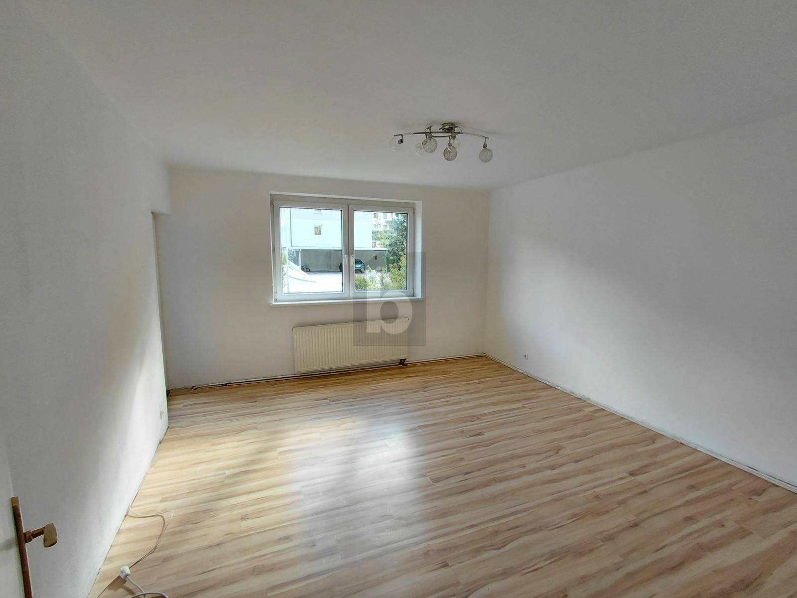 Leeres Zimmer mit Laminatboden und einem Fenster, das viel Tageslicht hereinlässt.