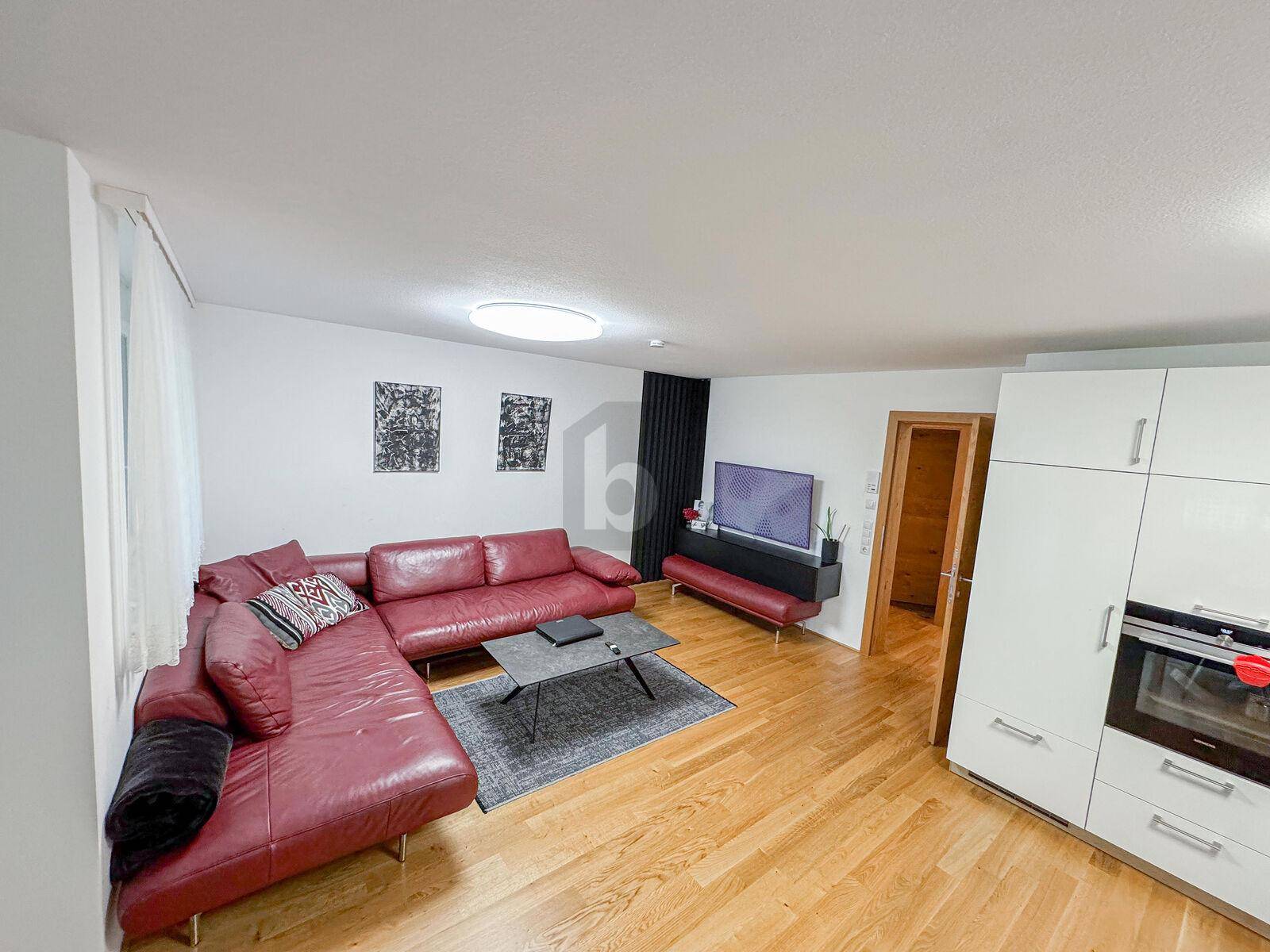 Gemütliches Wohnzimmer mit rotem Ledersofa, Holzboden und einem Fernseher, offen zum Küchenbereich.