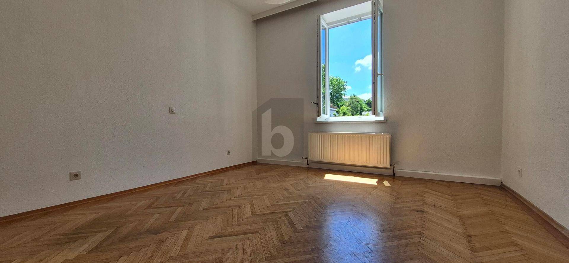 Leeres Zimmer mit Fischgrätparkett, weißen Wänden und Blick ins Grüne.