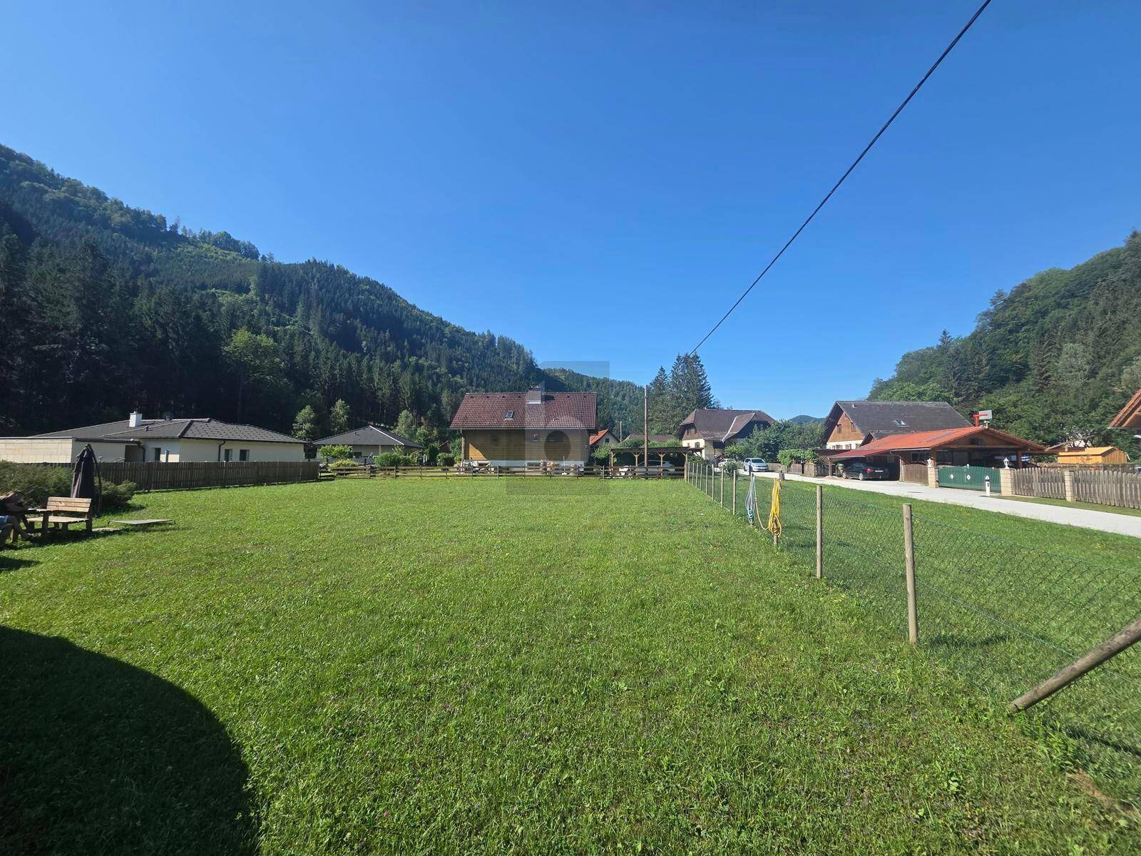 Grüne Wiese mit Zaun im Vordergrund, umgeben von Häusern und bewaldeten Hügeln unter klarem Himmel.