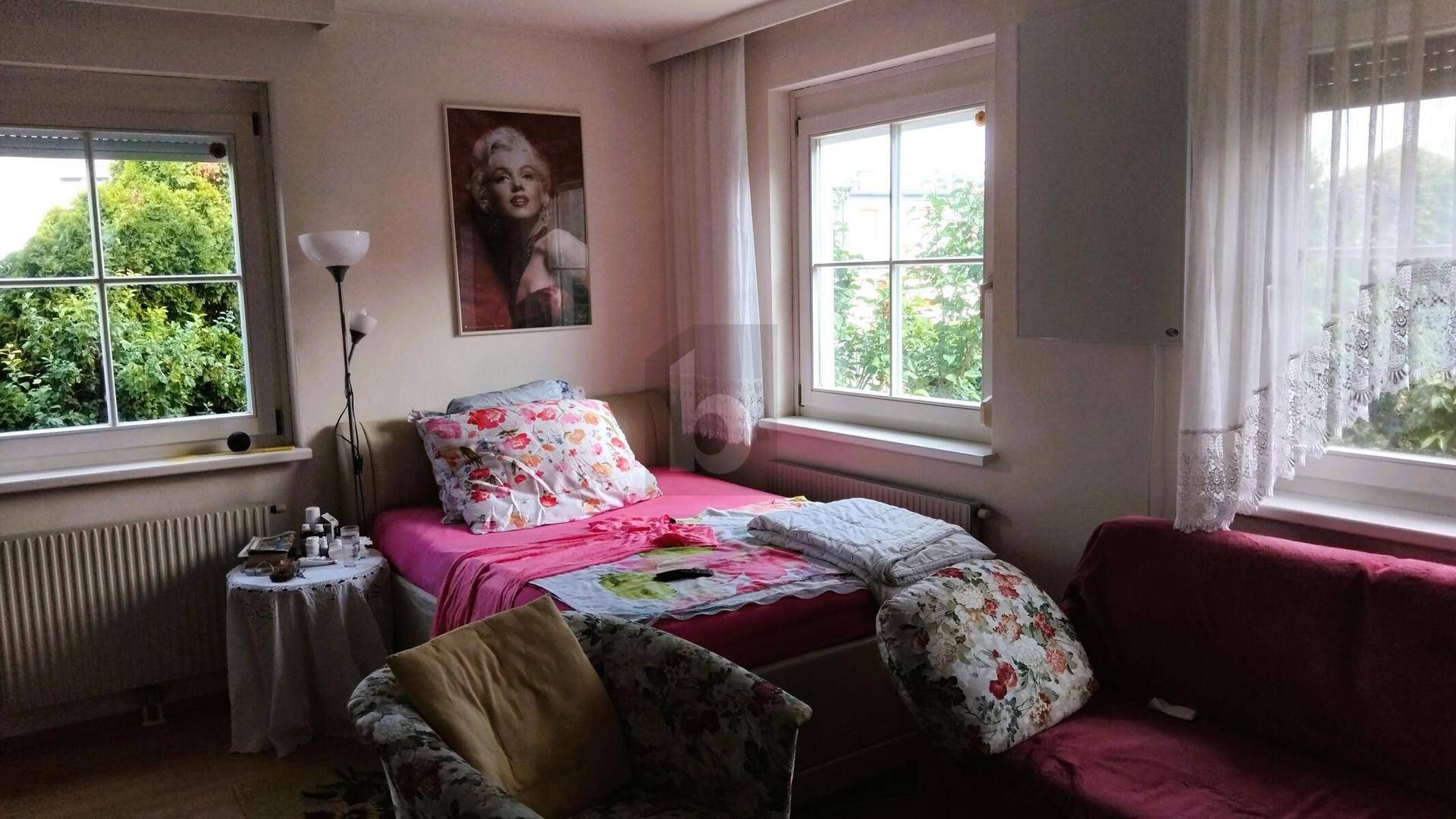 Gemütliches Schlafzimmer mit Einzelbett, Fenstern und einem Porträt an der Wand, umgeben von natürlichem Licht.
