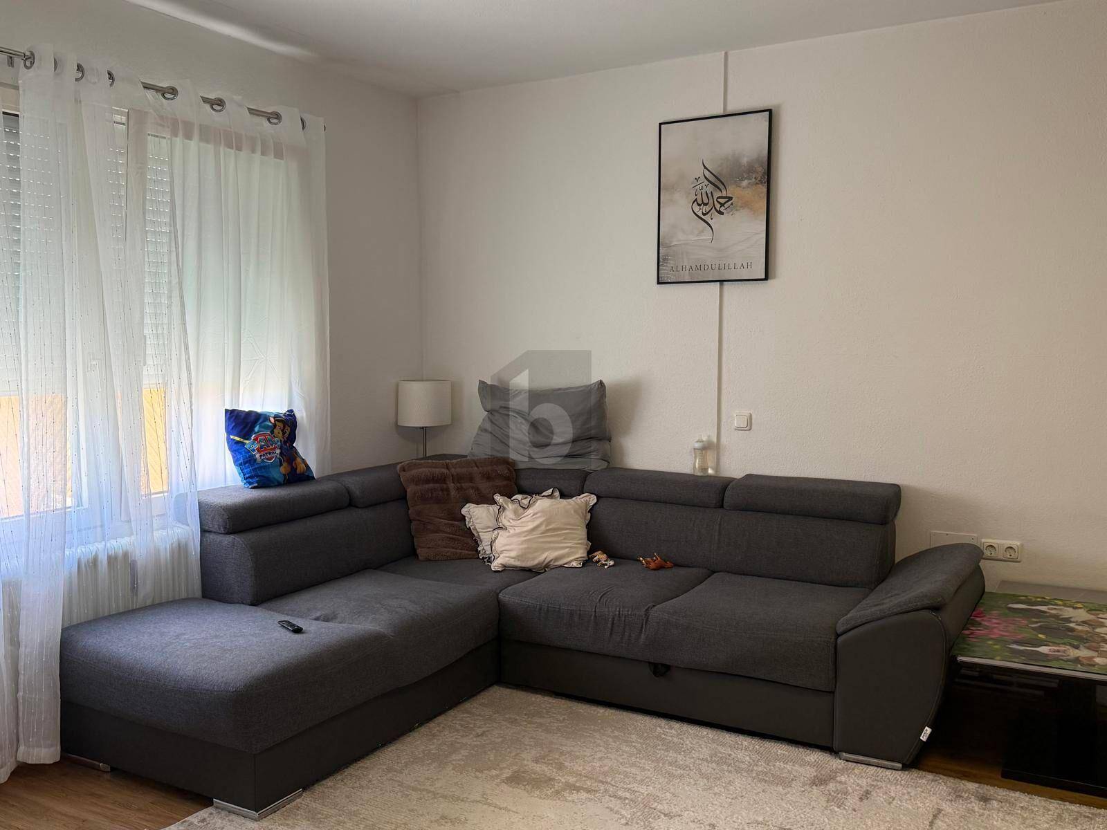 Gemütliches Wohnzimmer mit einem großen grauen Ecksofa und einem Fenster mit hellen Vorhängen.