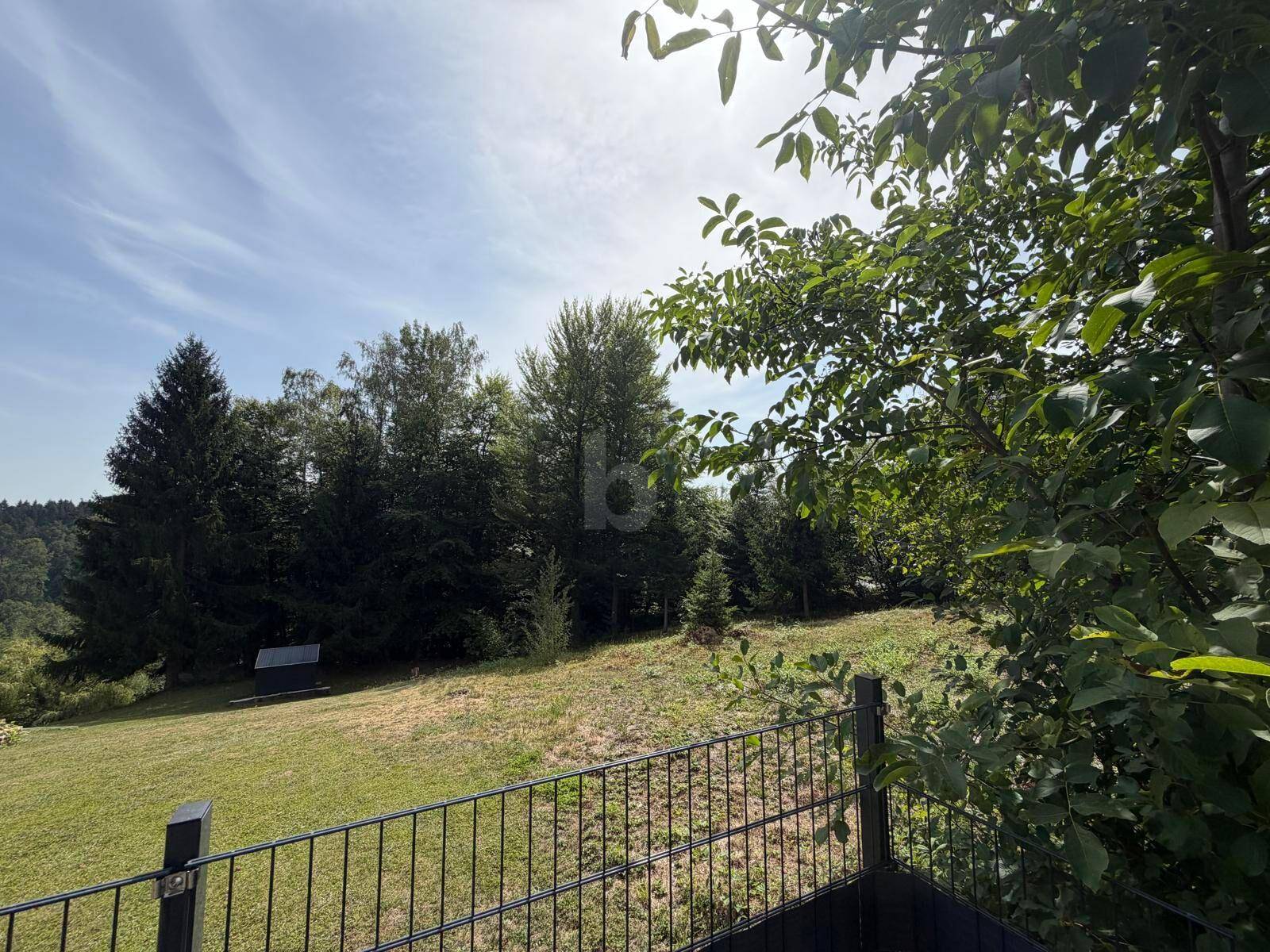 Natürliche Landschaft mit üppigem Baumbestand und einem Zaun im Vordergrund unter sonnigem Himmel.