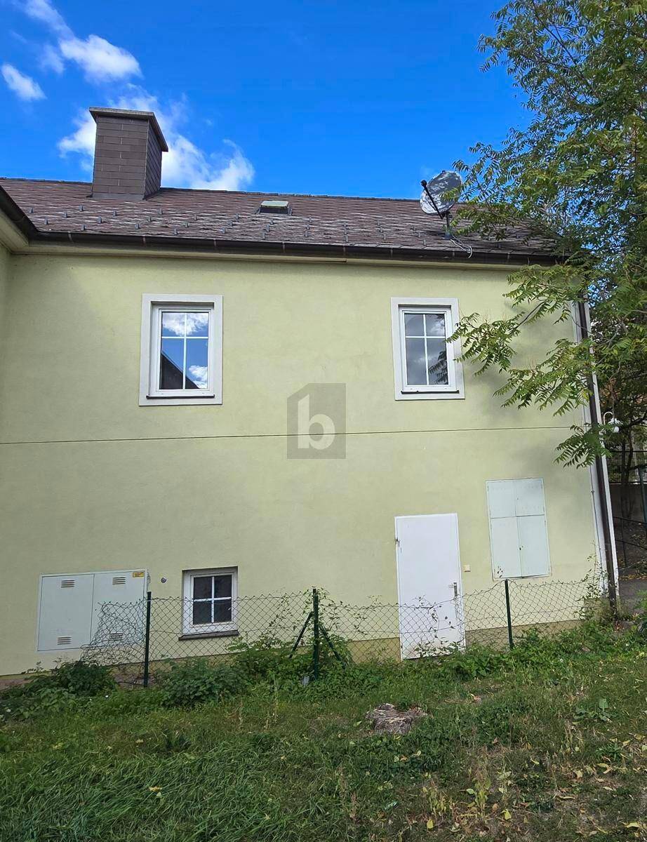 Seitenansicht des hellgelben Hauses mit Fenstern, Tür und Schornstein, umgeben von einem Zaun.