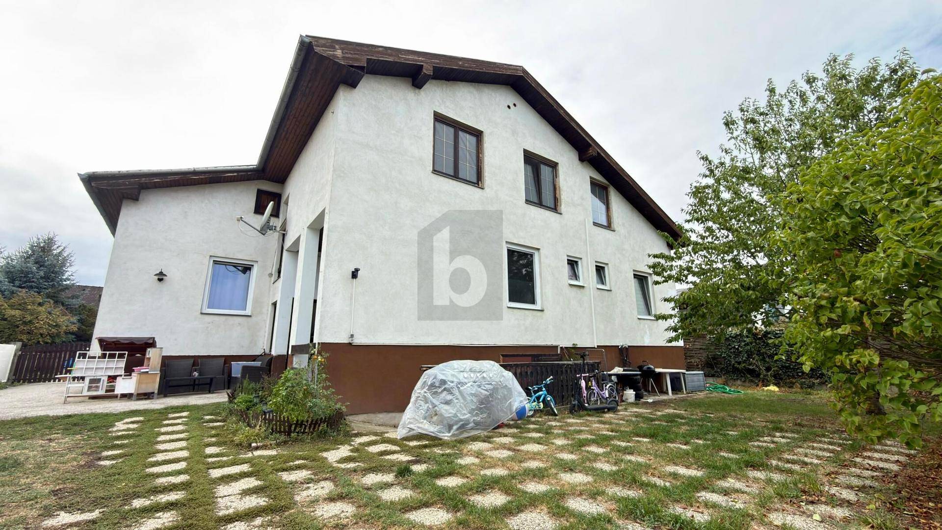 Rückseite des Hauses mit Gartenbereich, Rasenfläche und altem Baumbestand unter bewölktem Himmel.