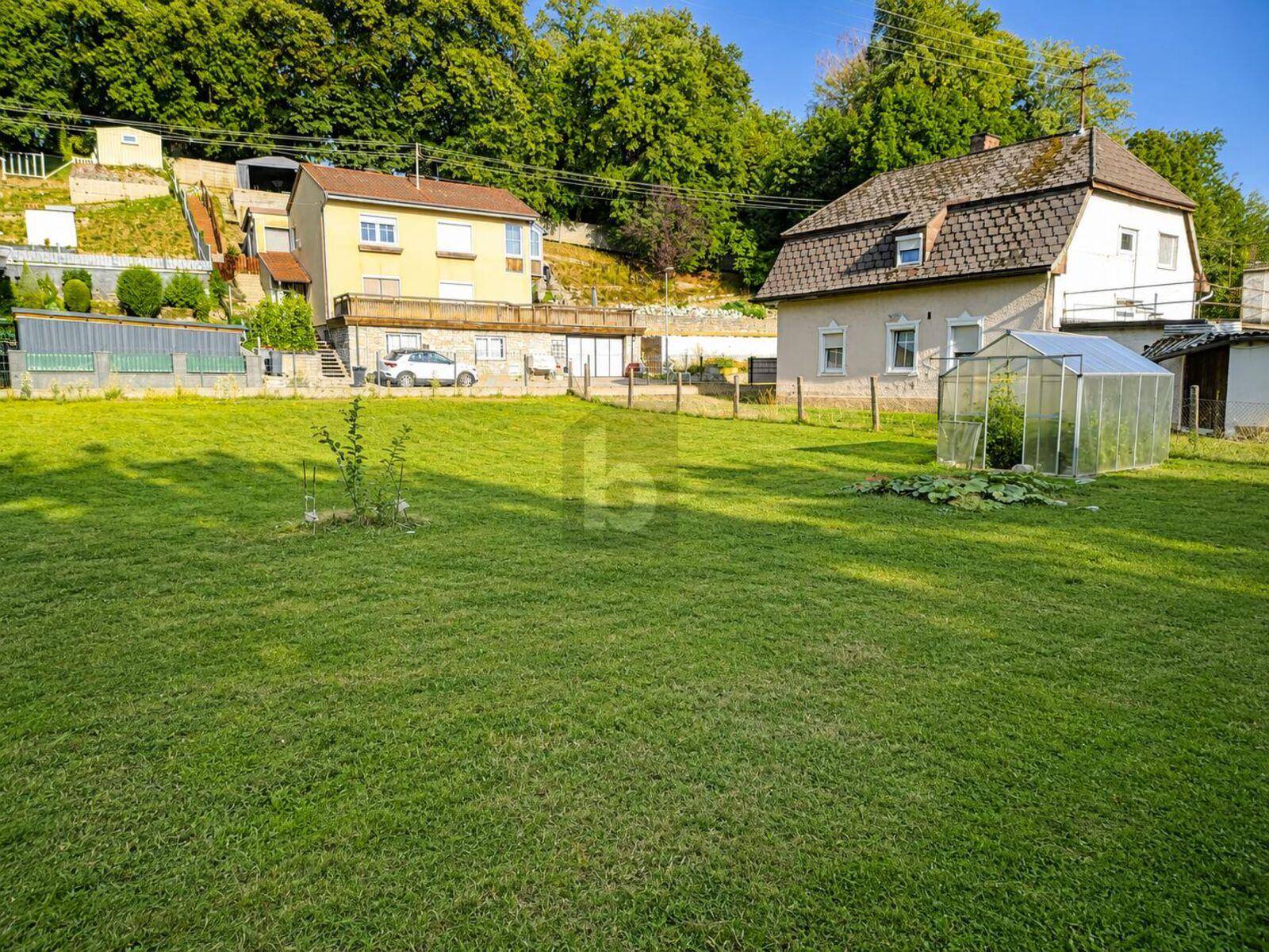 Große Rasenfläche mit Gewächshaus und Häusern am Hang, umgeben von grüner Landschaft.
