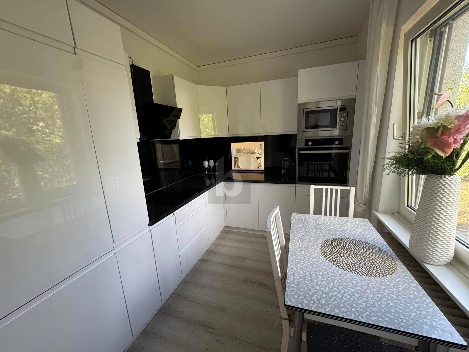 Moderne weiße Einbauküche mit schwarzen Arbeitsplatten, integrierten Geräten und Essbereich am Fenster.