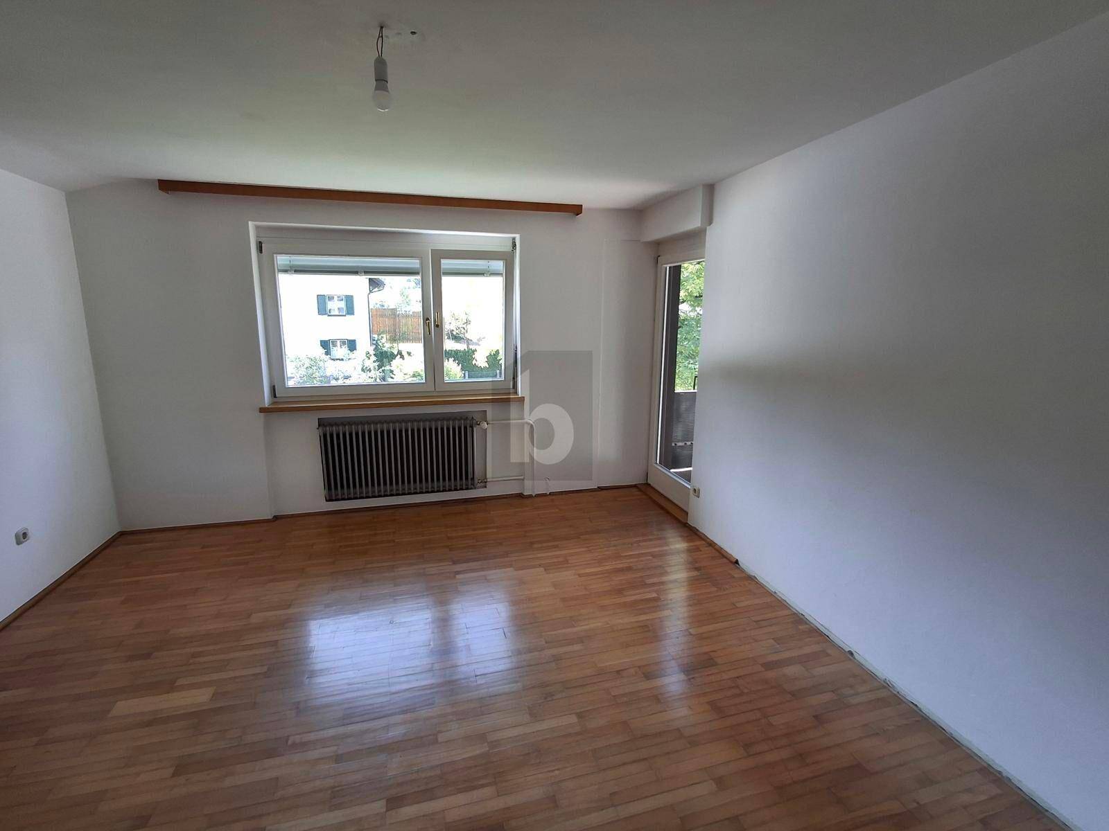Leeres Zimmer mit Parkettboden, weiß gestrichenen Wänden, Fenster und Zugang zu einem Außenbereich.