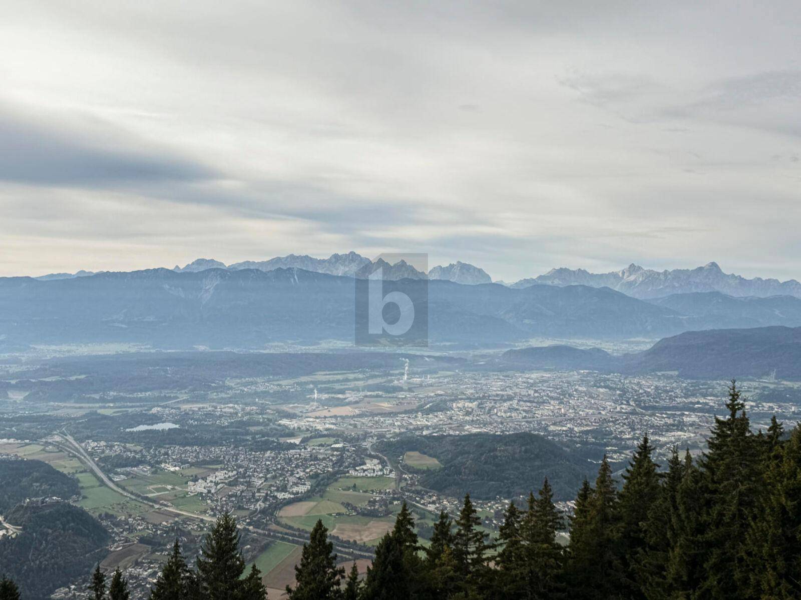 Panoramablick über eine Stadt und weite Berglandschaft unter bewölktem Himmel.