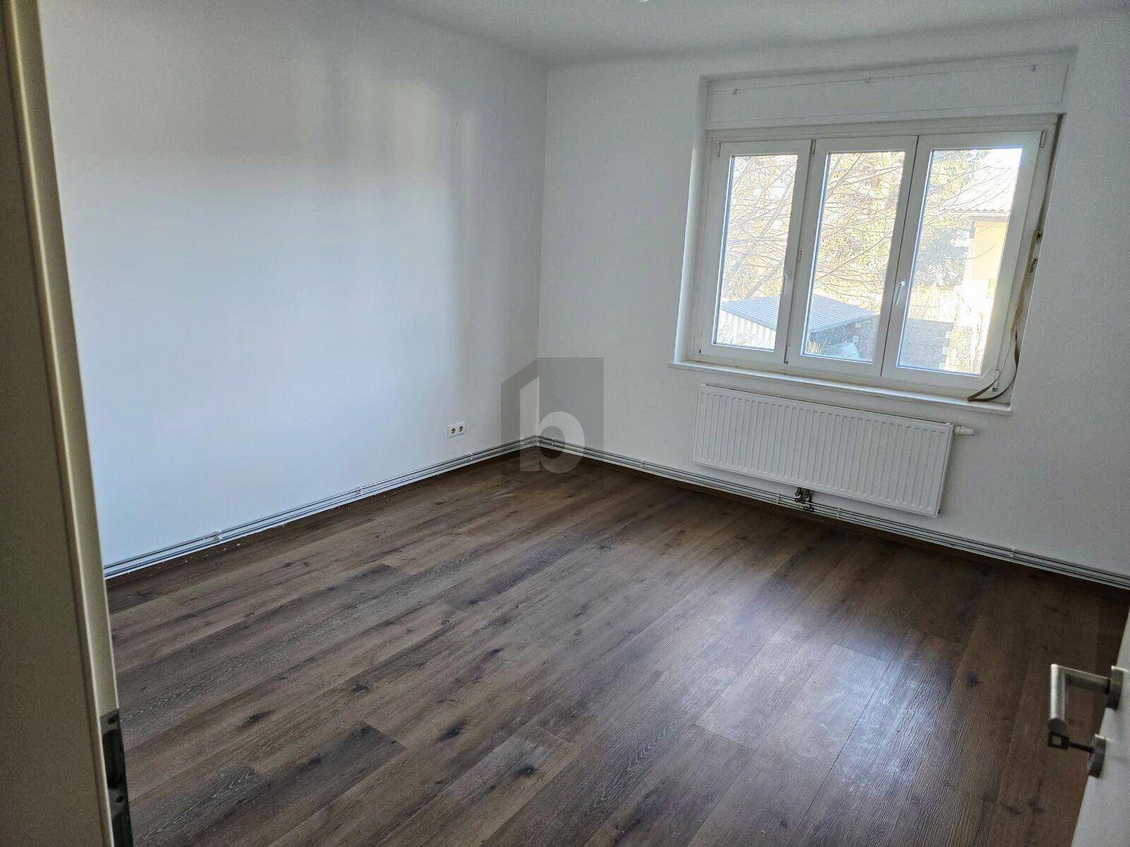 Schlafzimmer mit Laminatboden, weißen Wänden und einem großen Fenster mit Rollladen.