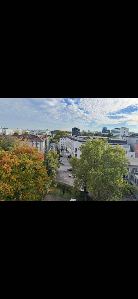 Weitläufiger Blick über eine städtische Umgebung mit viel Grün und Gebäuden unter blauem Himmel.