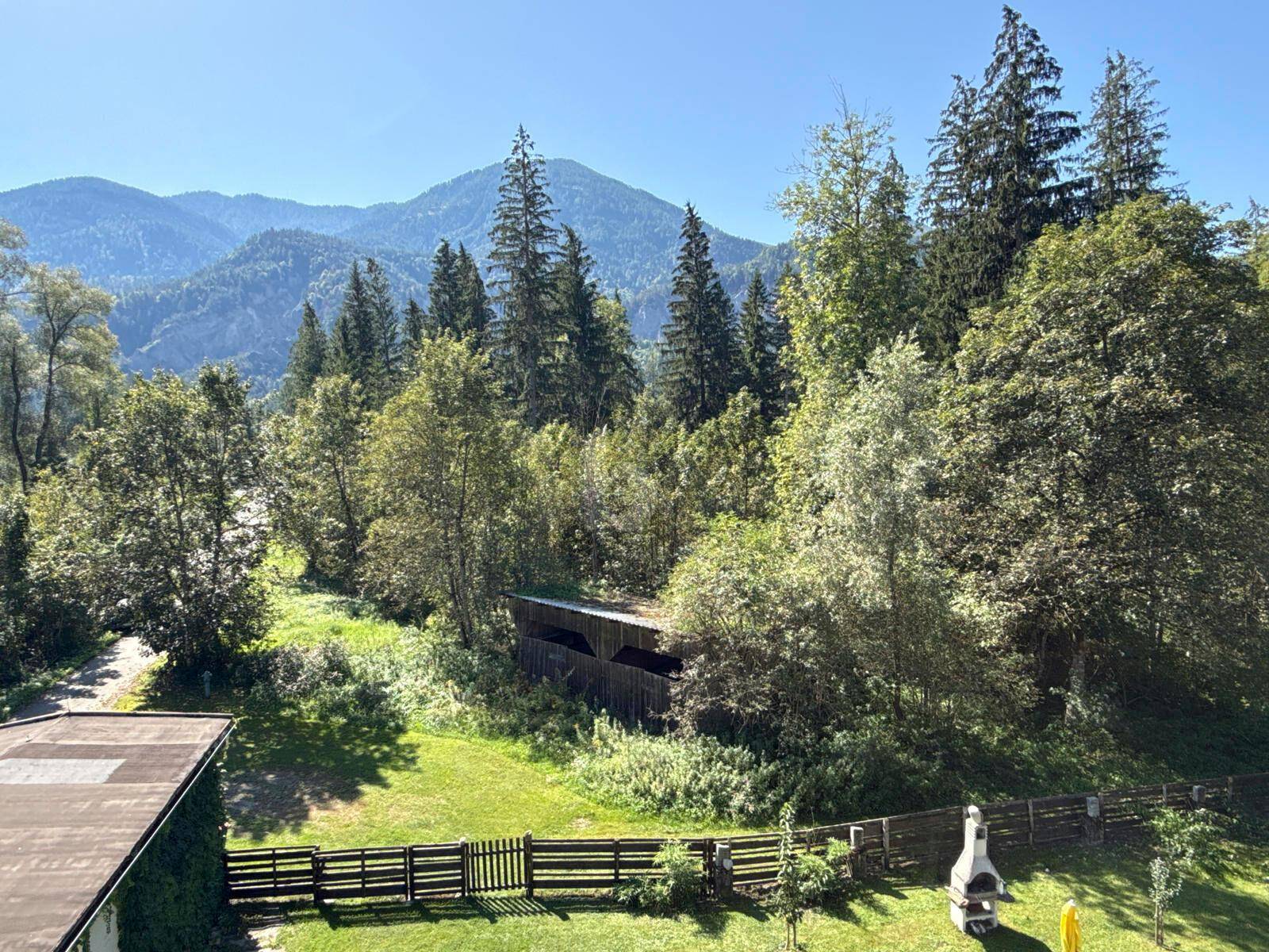 Malerischer Blick auf Berge und grüne Wälder von einem Gartenbereich mit Grill und Zaun.