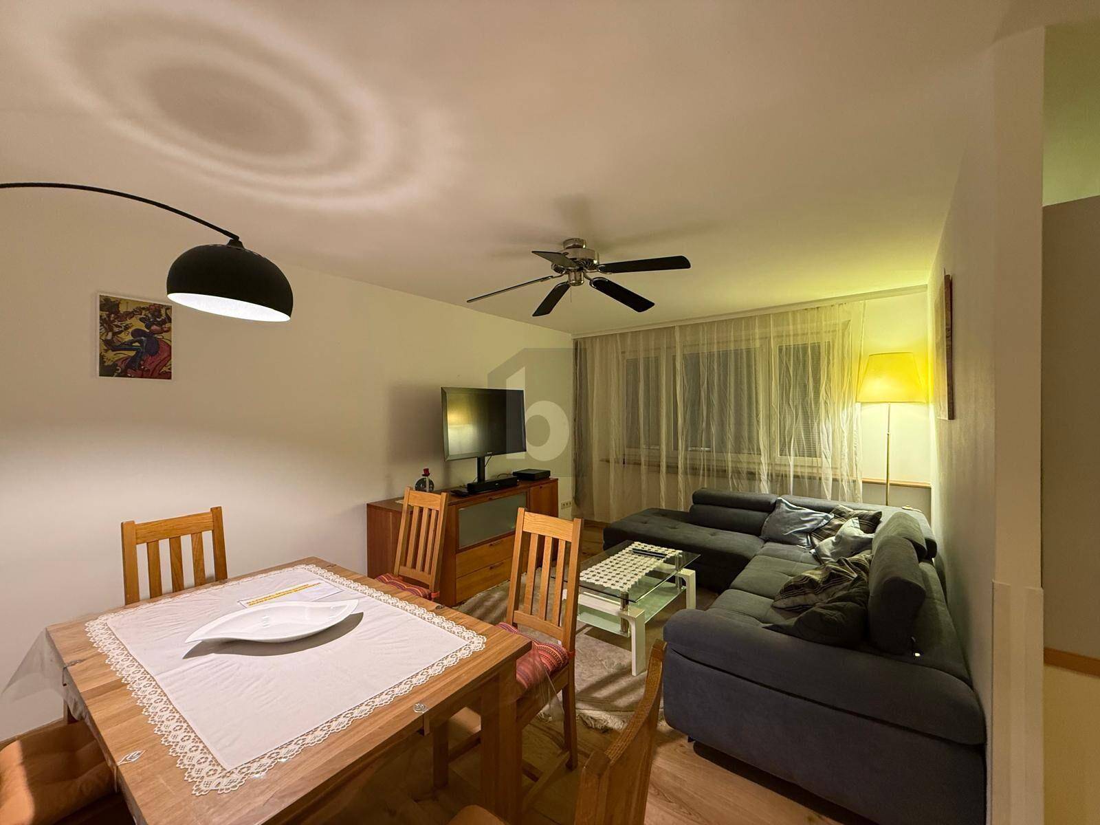 Gemütlicher Wohn- und Essbereich mit Holztisch, Ecksofa, Fernseher und einem Deckenventilator.