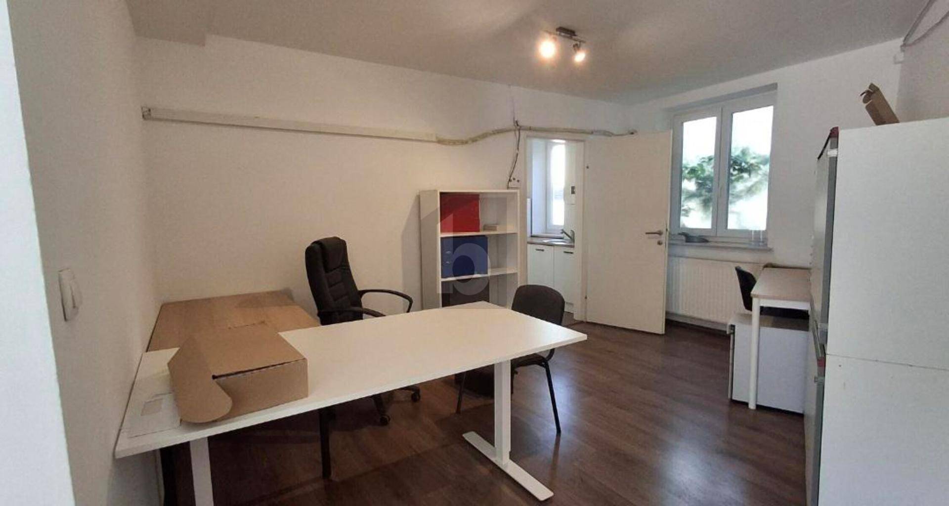 Funktionales Büro mit Schreibtisch, Bürostuhl und Fenster, angrenzend an einen kleinen Küchenbereich.