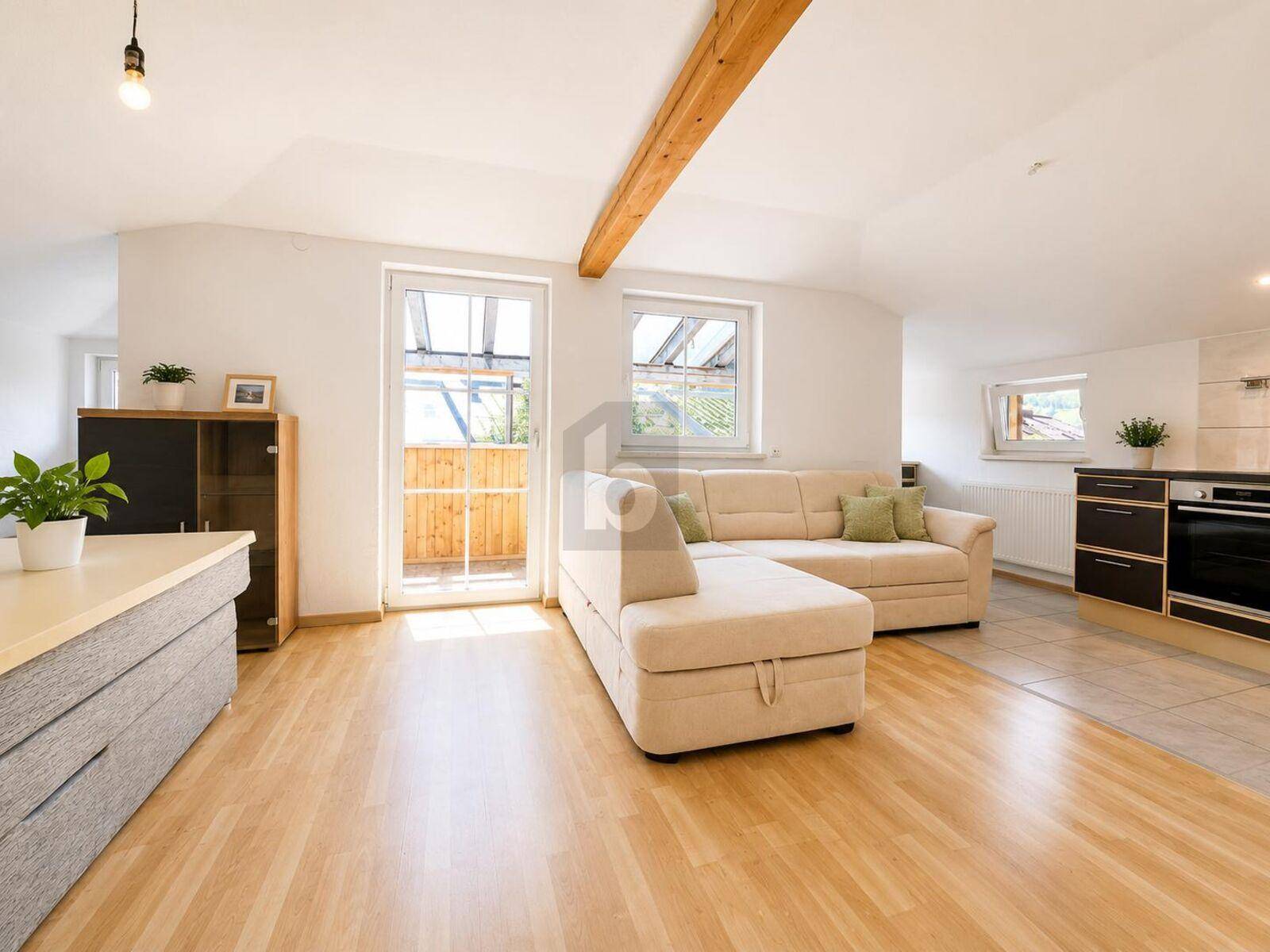 Heller Wohnbereich mit beigem Sofa, Holzboden, sichtbarem Deckenbalken und Balkonzugang.