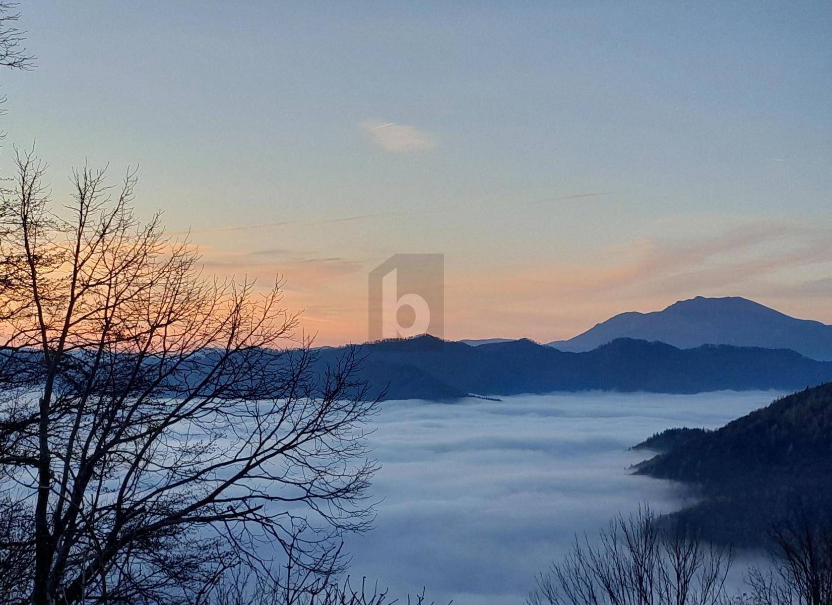 Malerische Berglandschaft bei Sonnenuntergang mit Nebel in den Tälern und kahlen Bäumen im Vordergrund.