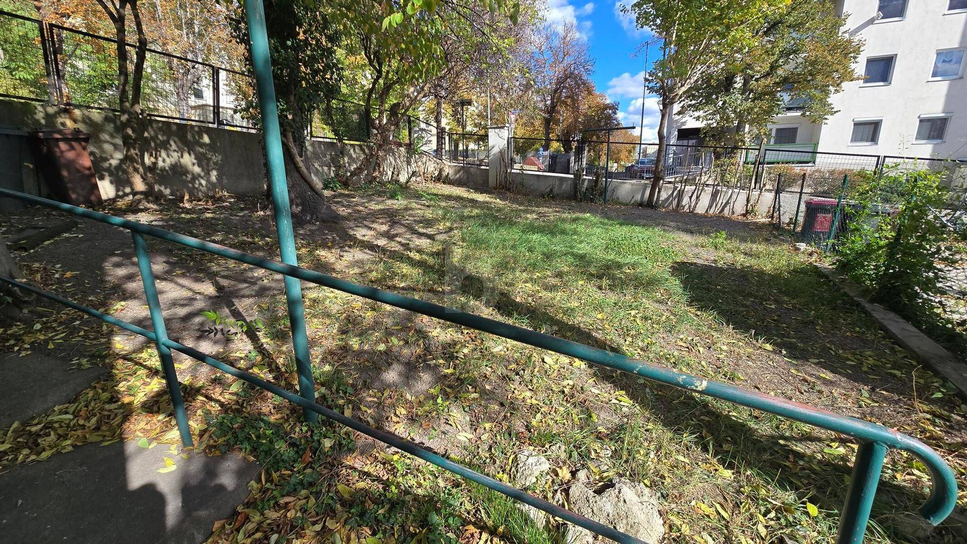 Außenbereich mit grünem Handlauf, Grasfläche und Bäumen, im Hintergrund ein Gebäude.
