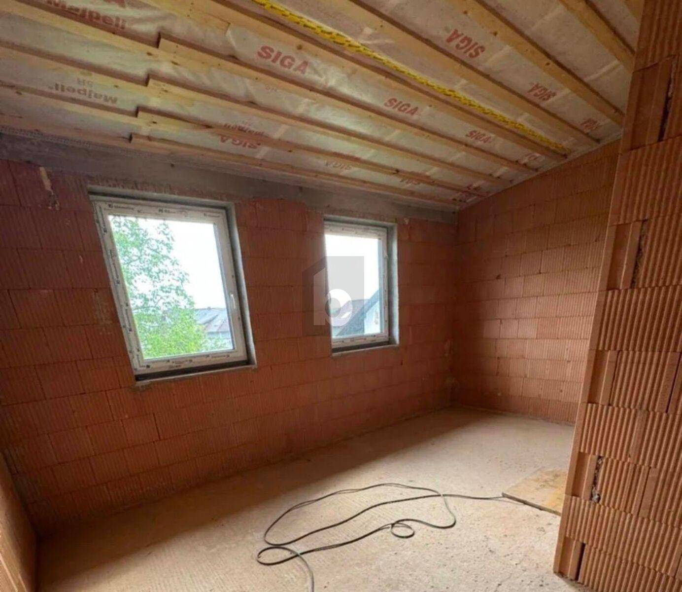 Unfertiges Zimmer im Obergeschoss mit Ziegelwänden, Holzbalkendecke und zwei Fenstern, bereit zum Ausbau.