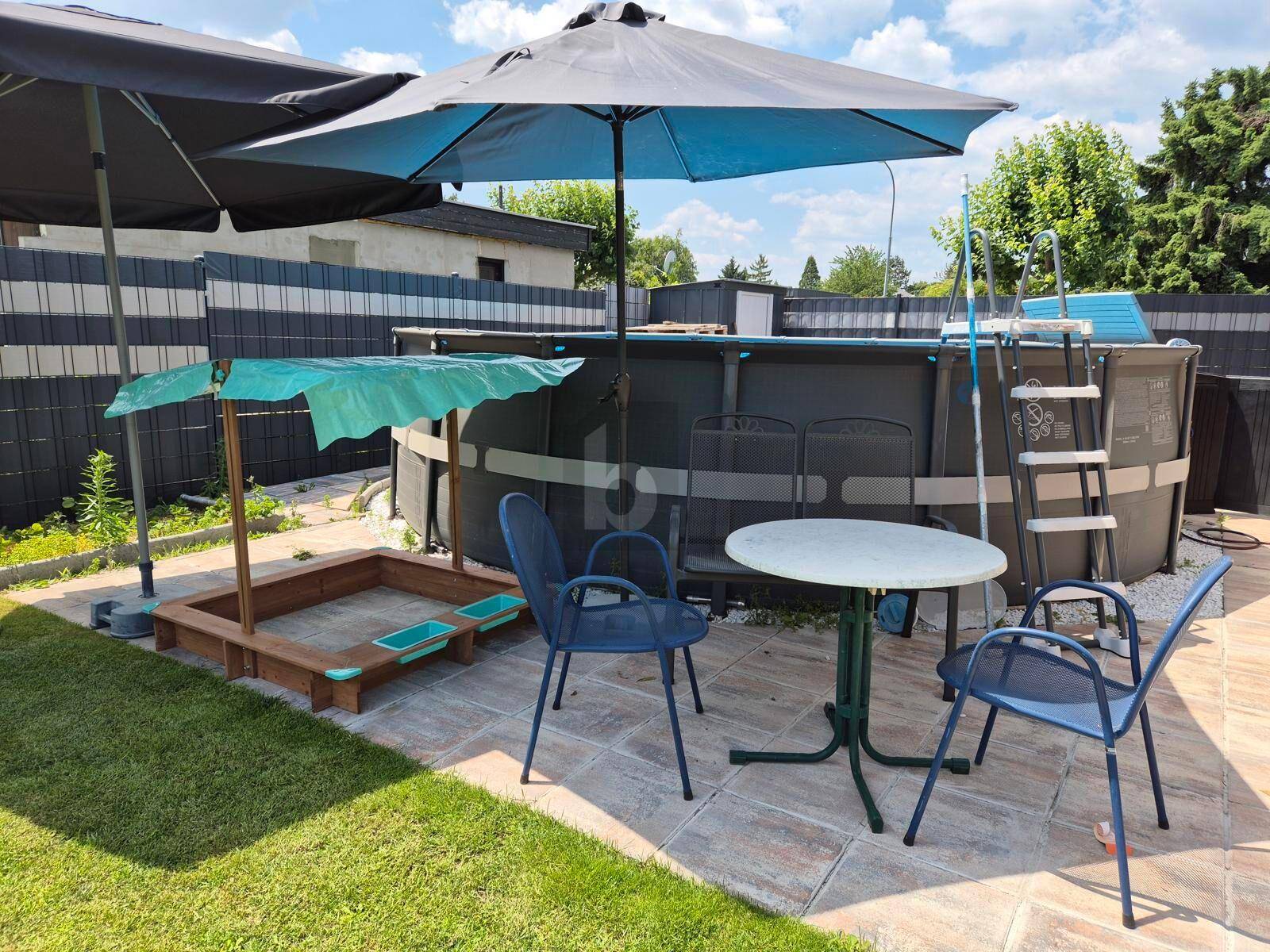 Gartenbereich mit großem Aufstellpool, Sandkasten und Sitzgelegenheiten unter Sonnenschirmen.