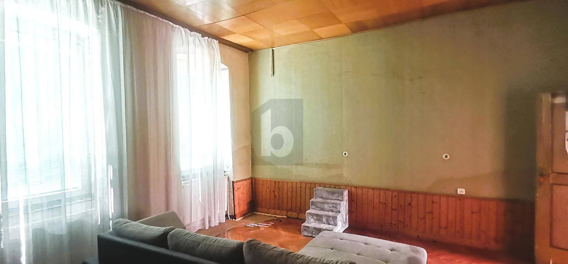 Wohnzimmer mit Sofa, Holzdecke, Holzvertäfelung und großen Fenstern mit Vorhängen.