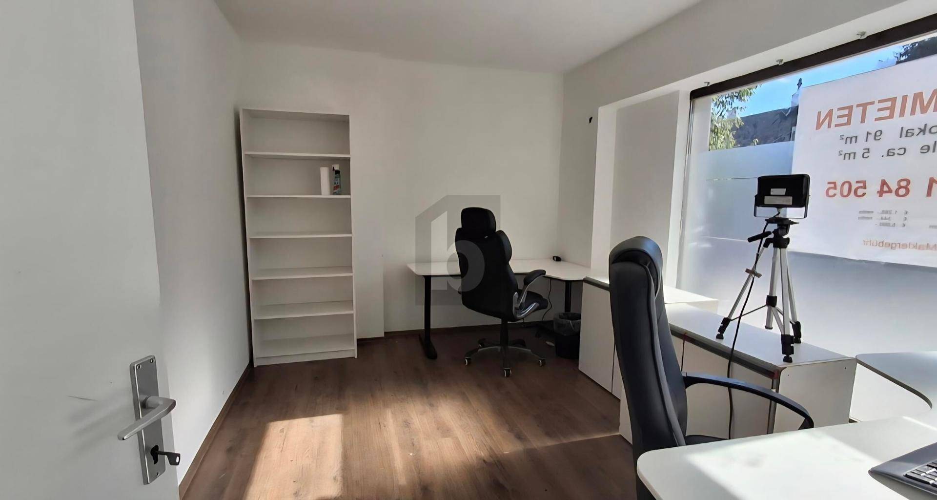 Büro mit Schreibtisch, Bürostuhl und Regal, beleuchtet durch ein großes Fenster mit einem "Zu vermieten"-Schild.