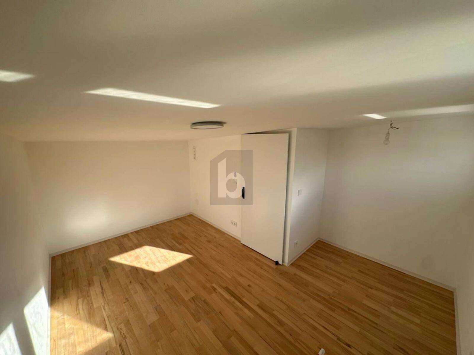Leerer Raum mit hellem Parkettboden und viel Tageslicht durch ein Fenster.