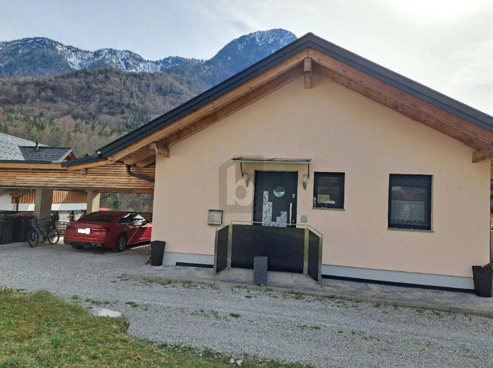 Hausfront mit Carport und Zufahrt, umgeben von einer beeindruckenden Berglandschaft.