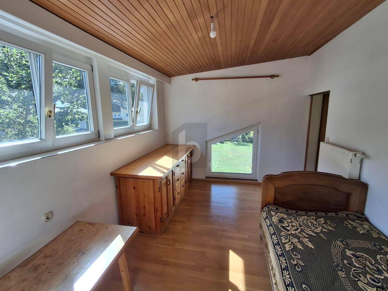 Helles Schlafzimmer mit Holzboden, Holzdecke, einem Bett und Blick ins Grüne durch mehrere Fenster.