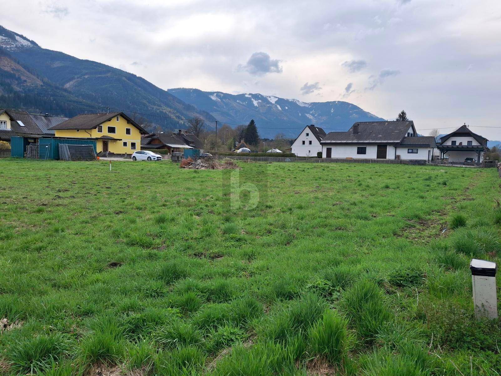 Weitläufige grüne Wiese mit umliegenden Häusern und schneebedeckten Bergen im Hintergrund.