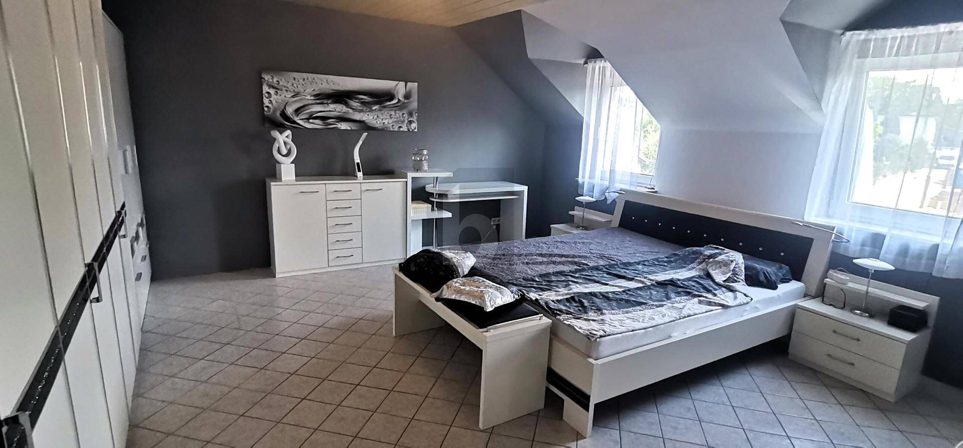 Schlafzimmer mit Dachschräge, Fliesenboden und modernem Bett in Grautönen.
