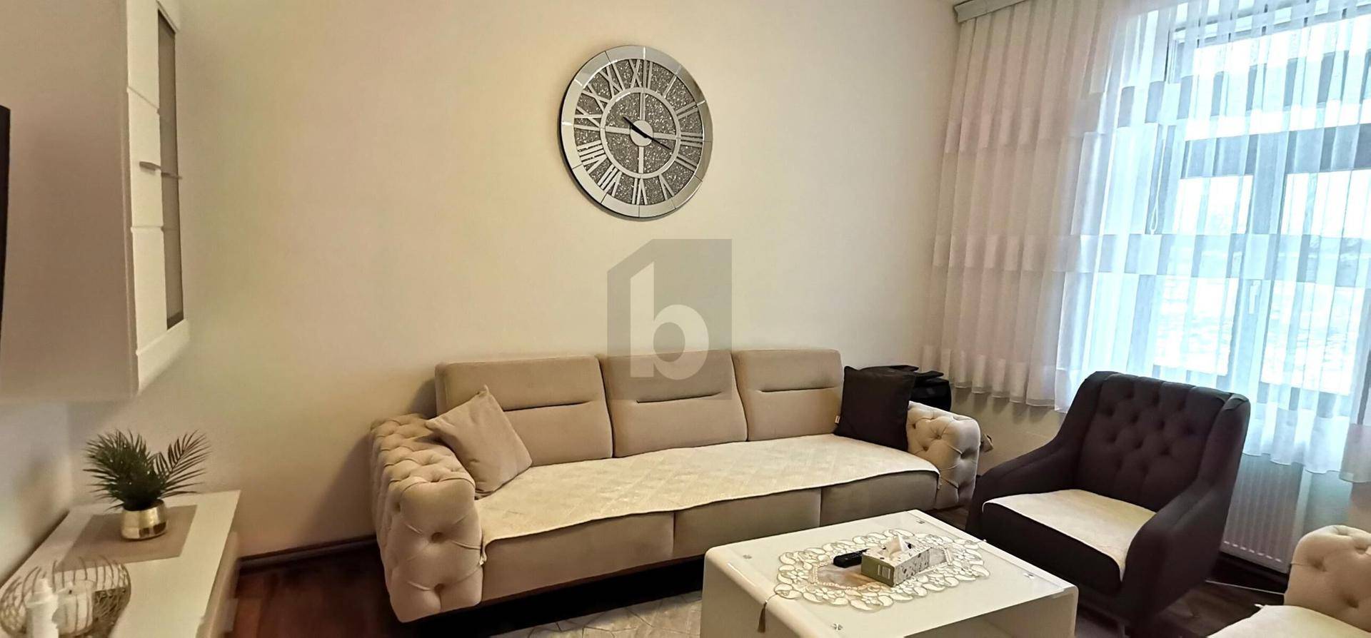 Gemütliches Wohnzimmer mit beigem Sofa, großem Wanduhr und Vorhängen am Fenster.