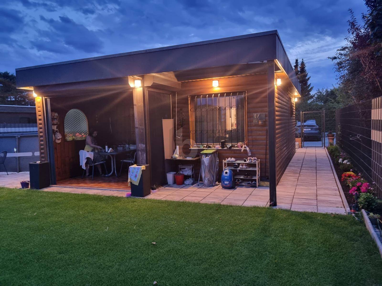 Beleuchtetes Gartenhaus aus Holz am Abend mit Terrasse und Blick auf den Rasenbereich.